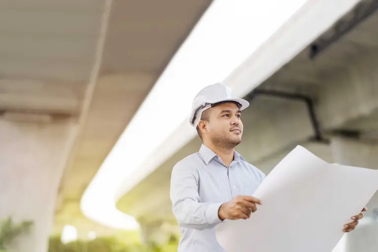 About us. Planner under concrete bridge with plans in hand. Planner wears white helmet.